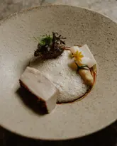 A high-quality, professional food photograph showcasing a refined starter dish, likely crab-based, featuring delicate foam, fresh herbs, and edible flowers on a textured ceramic plate. The image highlights the meticulous preparation and artistic plating typical of a fine dining establishment.
