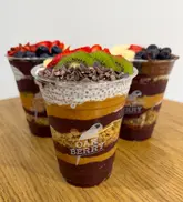 A clean, product-focused shot of three Oakberry açaí bowls arranged on a wooden table against a plain white background. The focus is sharp on the front bowl, highlighting the layers of açaí, nut butter, chia pudding, and granola, topped with fresh fruit and cacao nibs.