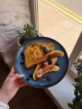 A top-down, handheld shot of a plate of French toast topped with caramelized bananas and blueberries, captured near a window in a cafe setting.