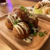 A close-up, high-quality shot of a serving of takoyaki on a wooden tray, topped with bonito flakes, mayonnaise, and green onions. The background is softly blurred, showing hints of other dishes and the restaurant environment.