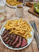 A top-down, casual shot of a steak and fries meal served in a restaurant setting, featuring a small jug of peppercorn sauce placed in the center of the table.