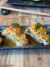 A close-up, high-quality shot of two pieces of nigiri sushi, featuring scallop and spicy crab, presented on a dark, textured plate against a wooden table background. The image effectively highlights the texture and freshness of the food, serving as a promotional post for a Japanese-Peruvian restaurant.