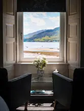 A high-quality, symmetrical shot of a hotel room interior looking out through a large sash window towards a scenic loch and mountain landscape. The composition frames the view with two armchairs and a small table with flowers in the foreground, creating an inviting and relaxing atmosphere.