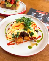 A bright, overhead food photography shot of a Brisket Benedict dish, featuring vibrant green and red oil drizzles, served at a cafe. The image is well-composed and highlights the texture and freshness of the food.