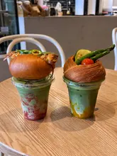 A photo featuring two drinks topped with food items—a prawn roll and a vegetable danish—sitting on a wooden table in a cafe setting.