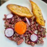 A close-up, top-down shot of a steak tartare dish served with toasted bread and radish slices, featuring a vibrant egg yolk as the focal point.