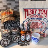A product haul photo featuring various Tubby Toms branded sauces, rubs, a hat, and a mug, arranged on a kitchen counter.