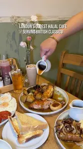 A high-angle, overhead shot of a table spread at a cafe, featuring a hand pouring gravy over a roast chicken dish, surrounded by various breakfast and lunch plates.