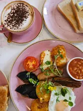 A top-down, flat-lay photograph of a breakfast plate featuring grilled halloumi, mushrooms, a roasted tomato, baked beans, and a fried egg, served on a pink plate alongside a cappuccino and toast.