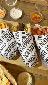 A handheld food haul video showcasing a variety of Greek takeaway dishes, including gyros, loaded fries, dips, and baklava, presented on a table for a group meal.