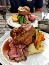 A high-quality, appetizing close-up shot of a Sunday roast dinner served outdoors. The image effectively uses natural light and a shallow depth of field to highlight the textures of the beef and the impressive size of the Yorkshire pudding, creating a strong desire to visit.