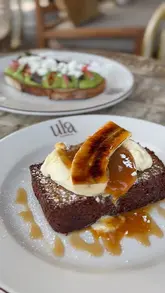 A high-quality, well-composed shot of a brunch spread featuring banana bread with cream and syrup in the foreground and avocado toast in the background. The lighting is soft and natural, highlighting the textures of the food effectively.