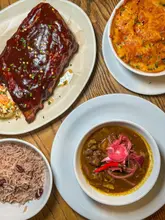 A top-down flat lay photograph of a Caribbean-style meal featuring BBQ ribs, mac and cheese, curry goat, and rice and peas on a wooden table.