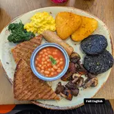 An overhead, flat-lay shot of a vegan Full English breakfast served on a speckled ceramic plate. The plate includes scrambled tofu or eggs, hash browns, black pudding, sautéed spinach, mushrooms, baked beans in a small bowl, and toast.