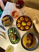 An overhead flat-lay shot of a restaurant table featuring a selection of small plates including steak, prawns, broccoli, and croquettes, accompanied by drinks.