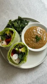 A clean, top-down photograph of homemade summer rolls served with a peanut dipping sauce. The rolls are filled with prawns, avocado, peppers, and cabbage, presented on a simple white plate against a textured white background, showcasing fresh and vibrant ingredients.