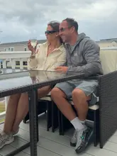 A lifestyle photograph of a couple sitting on a deck at a holiday resort, holding glasses of wine. The image captures a casual, relaxed moment during a family trip.