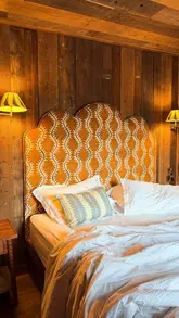 A warm, inviting shot of a luxury lodge bedroom featuring a distinctive scalloped headboard against rustic wood-paneled walls. The lighting is soft and atmospheric, emphasizing the cozy, high-end nature of the accommodation.