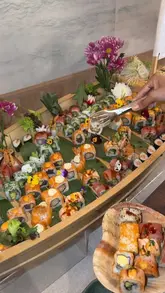 A promotional shot of a sushi boat at a new restaurant opening, featuring a variety of sushi rolls presented on a wooden boat with floral garnishes.