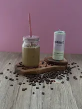 A product shot featuring a can of Grind Oat Vanilla Latte alongside a glass of the same drink, set against a plain pink background on a wooden surface.