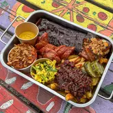 A top-down shot of a metal tray filled with a variety of BBQ meats, including ribs, sausage, and brisket, accompanied by sides of beans, coleslaw, and pickles, served on a brightly painted picnic table.
