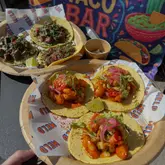 A vibrant, overhead shot of freshly prepared tacos served on branded paper plates at an outdoor event. The tacos are topped with fresh herbs, pickled onions, and lime, with a 'Taco Bar' banner visible in the background, creating a lively festival atmosphere.