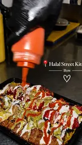 A close-up, top-down shot of a takeaway container being drizzled with sauce, showcasing a loaded meal of various meats, rice, and chips from a ghost kitchen.