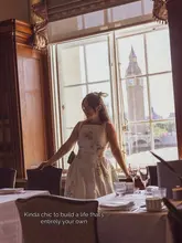 A lifestyle shot featuring a woman standing in a formal dining room with a prominent, iconic view of Big Ben and the River Thames through a large window, suggesting a high-end hotel or restaurant setting.