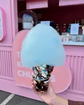 A bright, eye-catching shot of a dessert cup featuring soft-serve ice cream, chocolate sauce, and a large cloud of blue candy floss, held in front of a branded pink kiosk. The image effectively highlights the 'Instagrammable' nature of the product, using vibrant colours and a clean, focused composition to draw the viewer's attention to the treat.