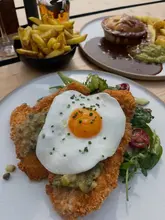 A casual, overhead shot of a pub lunch featuring a chicken schnitzel topped with a fried egg and a steak and ale pie with chips, taken in an outdoor setting.