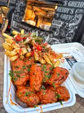 A close-up shot of a takeaway container filled with buffalo-style chicken wings and loaded fries, held in front of a branded food truck. The image captures the messy, indulgent nature of the food, highlighting the sauce and toppings.