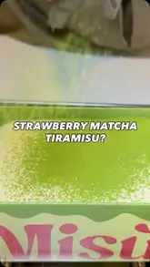 A close-up, high-quality shot of matcha powder being dusted over a strawberry matcha tiramisu in a branded takeaway box. The video captures the preparation/finishing touch, highlighting the texture and freshness of the dessert.