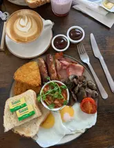 A top-down, flat-lay shot of a classic British fry-up breakfast accompanied by a latte. The image captures the components of the meal clearly on a wooden table surface.