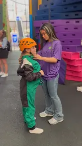 A clear, functional shot taken at Clip 'n Climb at The O2, showing a staff member helping a child put on a climbing harness. The background features the colourful climbing wall structure, establishing the venue's activity-focused environment.
