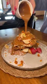 A close-up, high-angle shot of a stack of pancakes being drizzled with a caramel-coloured sauce, likely at a cafe. The focus is on the texture of the pancakes and the sauce pour.