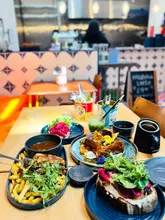 A high-angle, flat-lay shot of a brunch spread in a cafe, featuring a grilled toastie, salmon on rye, and a matcha drink. The image captures a vibrant, appetizing meal setting with a blurred background that suggests a busy, welcoming atmosphere.