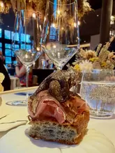 A close-up, high-quality shot of a sophisticated dish featuring bread topped with cured meat and shaved truffle, captured in a moody, upscale restaurant setting. The shallow depth of field draws the eye directly to the textures of the food, while the blurred wine glasses in the foreground add depth and context to the fine dining experience.