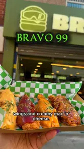 A close-up, handheld shot of a takeaway tray featuring four distinct styles of fried chicken wings, held in front of the 'Bravo 99' storefront. The image effectively showcases the 'messy' comfort food aesthetic, with the wings appearing saucy and well-prepared. The background is softly blurred, keeping the focus entirely on the food.