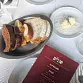 A high-quality, top-down flat-lay photograph of a table setting at BÔNE in Hoxton. The image features a basket of assorted breads and a small plate of parmesan butter, accompanied by the restaurant's menu. The lighting is bright and natural, creating an inviting and clean aesthetic.