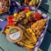 A top-down, 45-degree angle shot of a takeaway feast featuring fried chicken and fries, presented in branded packaging on an outdoor picnic table.