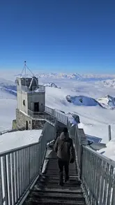 A short travel clip featuring a person walking down metal stairs at the Glacier Paradise viewing platform in Zermatt, Switzerland, followed by a panoramic view of the surrounding snow-covered Alps.