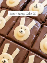 A top-down, instructional recipe video demonstrating how to create an Easter Bunny-themed chocolate traybake. The video uses clear text overlays to guide the viewer through the steps, from frosting the cake to assembling the chocolate button decorations.