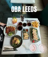 An overhead flat-lay shot of a table spread at Oba Leeds, showcasing a variety of Korean and Japanese dishes including sushi, spring rolls, fried chicken, fries, and cocktails.