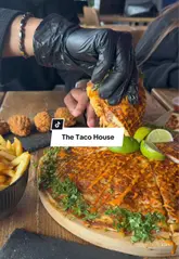 A short, punchy review of The Taco House, featuring close-up shots of a large taco pizza, crispy wings, and fries. The video captures the social dining experience with friends, focusing on the texture and appeal of the food.