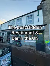 A static image of the exterior of 'The Secret Garden' in Pooley Bridge, featuring a shop-front entrance cluttered with retail items, overlaid with text claiming it is the 'best hidden restaurant & bar in the UK'.